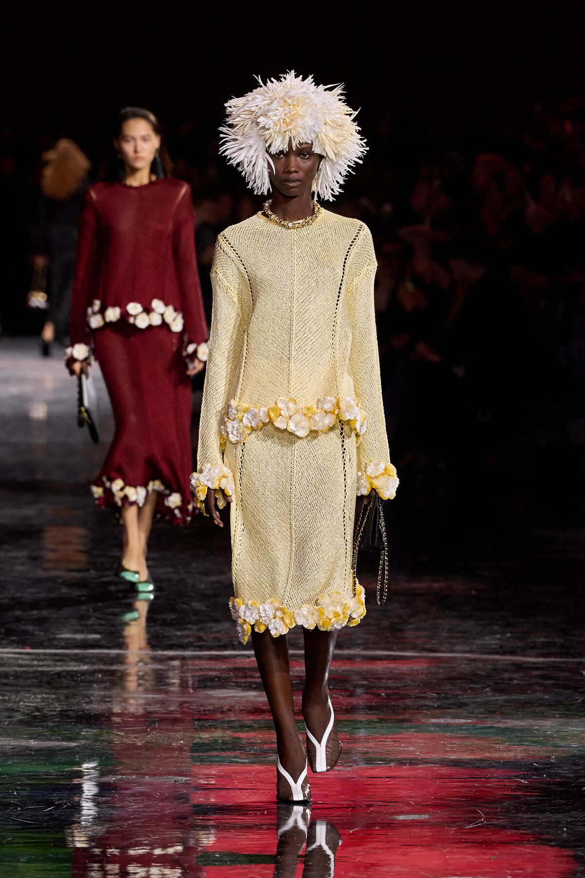 Model walking a runway in a pale yellow knit dress with white flower trim and a large white feather hat.