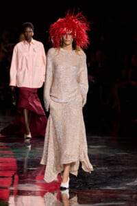 Model on a runway wearing a shimmering beige gown and a large red feathered hat, mid-stride during a fashion show.