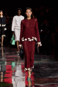 Model walking a burgundy knit gown with cream flower pom-poms along the hem and cuffs, on a glossy runway at a fashion show