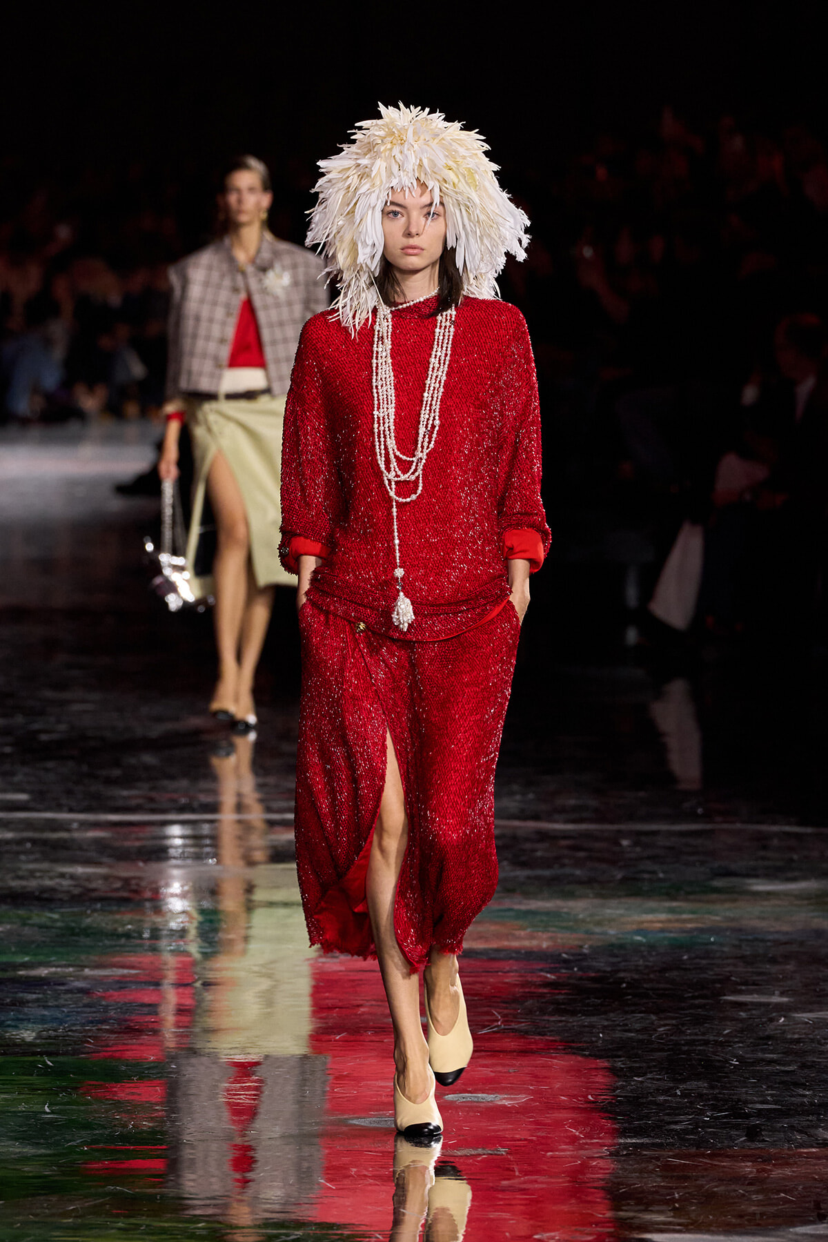 Model walks the runway in a red sequin outfit with a long pearl necklace and a dramatic white feathered hat.