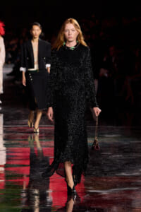 Model walking a glittering black sequin gown down the runway, holding a small burgundy purse with a chain strap.