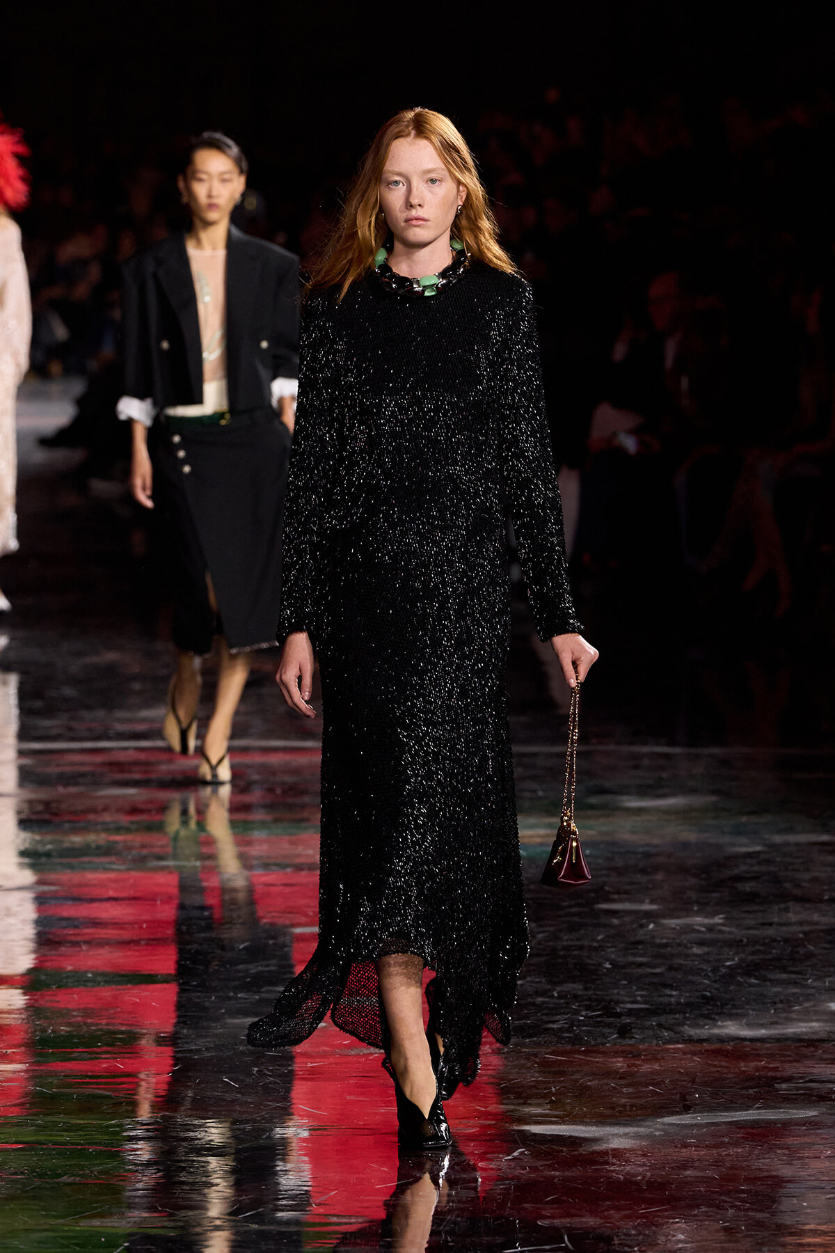 Model walking a glittering black sequin gown down the runway, holding a small burgundy purse with a chain strap.