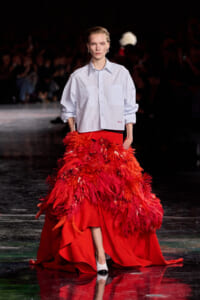 Runway model in a light blue button-down shirt with a voluminous red feathered gown and white shoes.