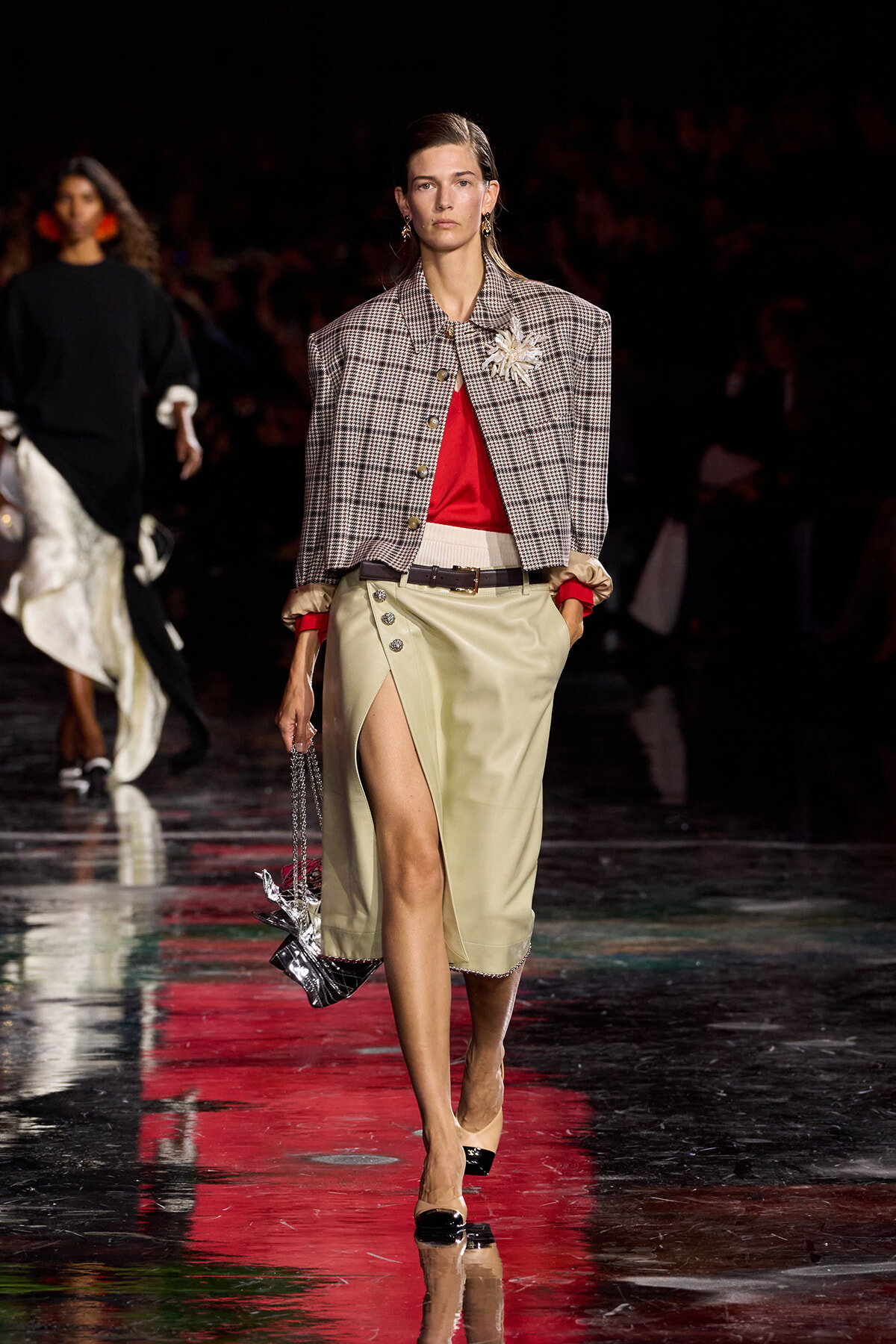 Model walking a fashion runway in a plaid cropped jacket over a red top, beige high-slit skirt, belt, and a metallic handbag with a decorative brooch at the shoulder.