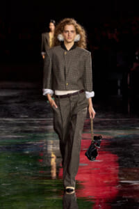 Model walks the runway in a brown checkered suit with rolled sleeves, white fluffy statement earrings, and a small black handbag on a gold chain.