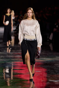 Model walks the runway in a cream satin blouse with two front pockets tucked into a black midi skirt with a front slit, accessorized with a beige belt and burgundy clutch.