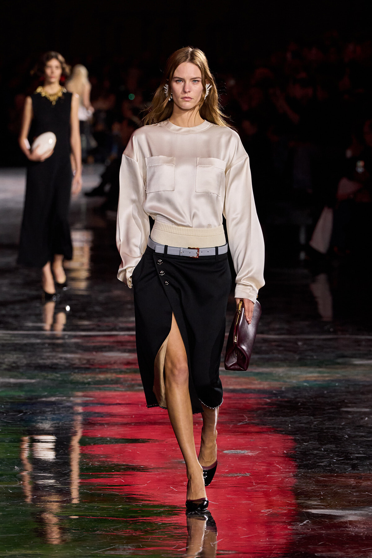 Model walks the runway in a cream satin blouse with two front pockets tucked into a black midi skirt with a front slit, accessorized with a beige belt and burgundy clutch.