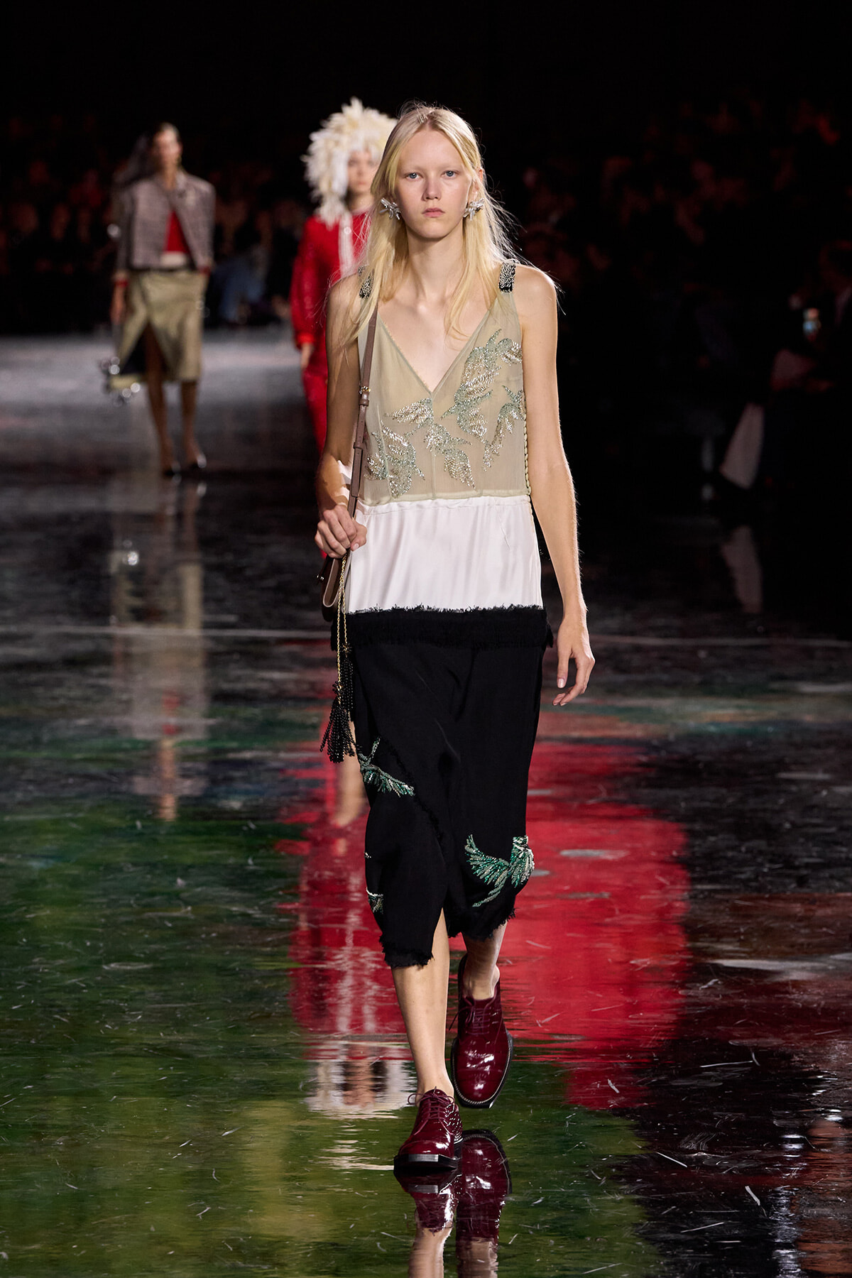 Model walking a fashion runway in a beige embroidered sleeveless top with a white and black fringed skirt and burgundy shoes; carries a small brown crossbody bag.