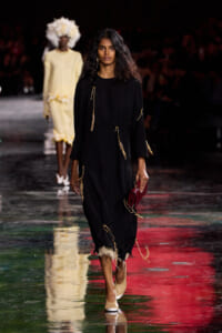 Model walks the runway in a loose black dress adorned with gold tassels, carrying a red clutch and beige heels on a glossy runway.