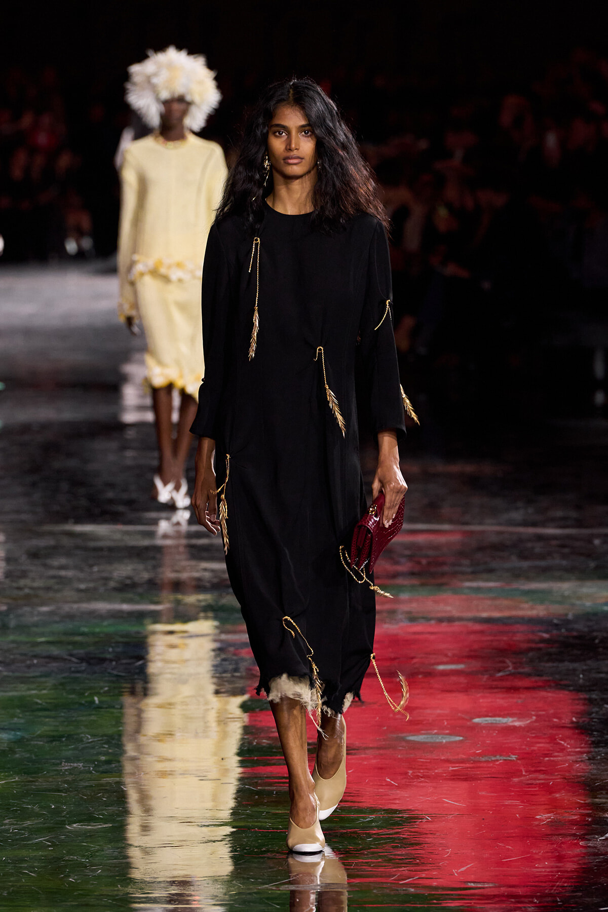 Model walks the runway in a loose black dress adorned with gold tassels, carrying a red clutch and beige heels on a glossy runway.