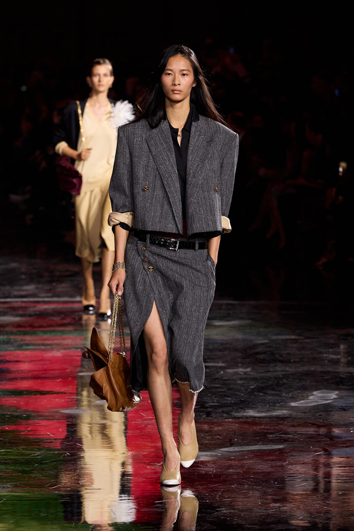 Model walking a fashion runway in a charcoal gray double-breasted suit with a high slit skirt and beige heels, carrying a brown handbag.
