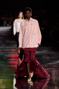 Model walks runway in oversized pale pink shirt with white corsage, burgundy high-low skirt, and two-tone heels carrying a black clutch.