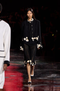 Model walking runway in a black textured suit with white floral appliqués at cuffs, hem, and collar, paired with sheer black pants and black‑white heels.