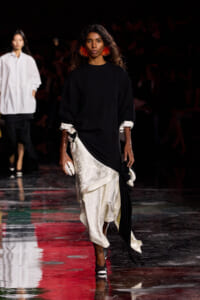 Model walking runway in black oversized top with white lace skirt and red fringe earrings, holding a white clutch.