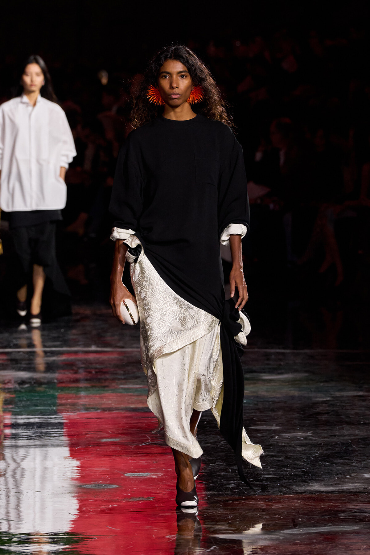 Model walking runway in black oversized top with white lace skirt and red fringe earrings, holding a white clutch.