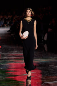 Model walking a runway in a sleeveless black jumpsuit, holding a white oval clutch and wearing a gold statement necklace with red earrings.