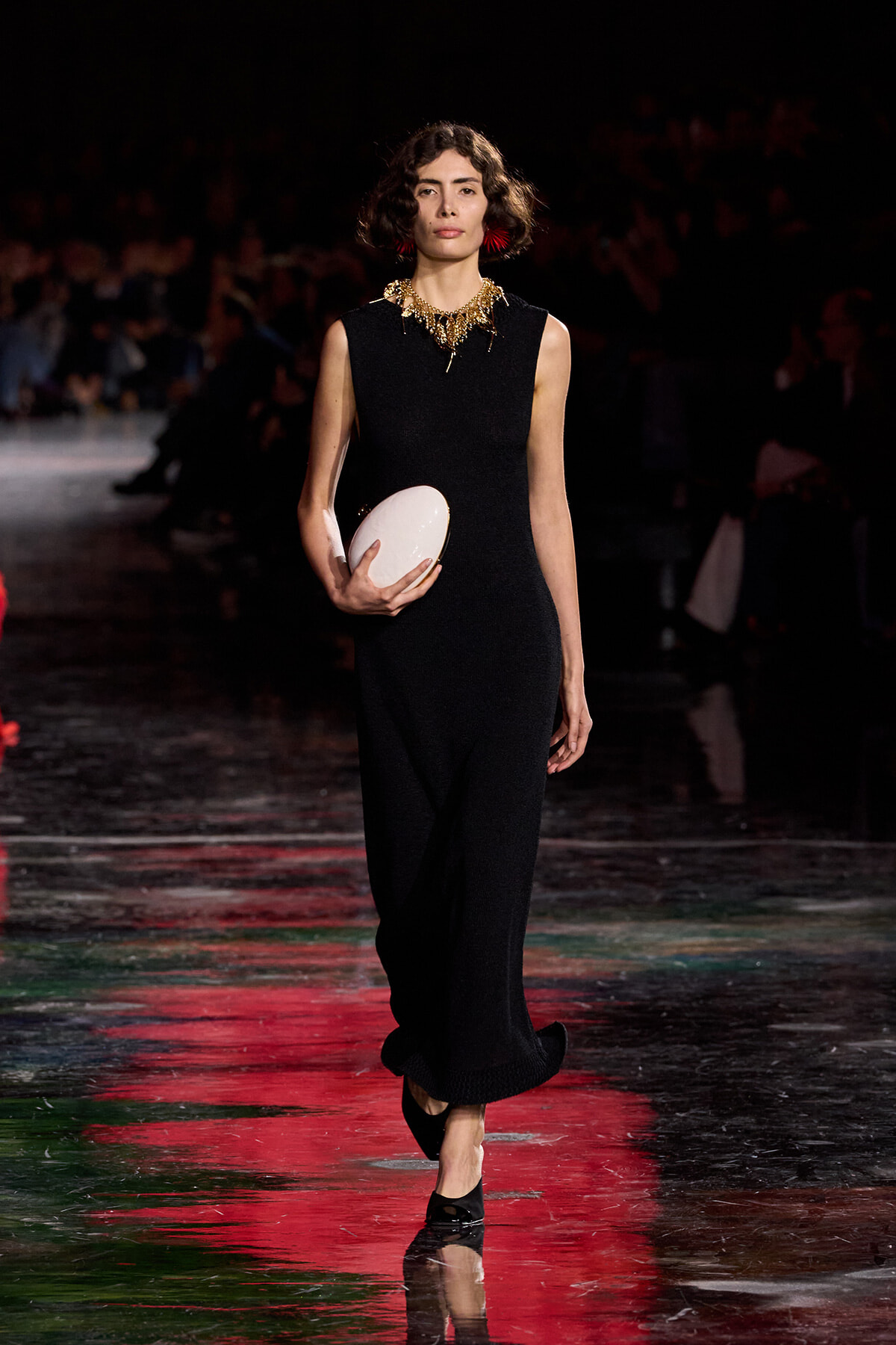 Model walking a runway in a sleeveless black jumpsuit, holding a white oval clutch and wearing a gold statement necklace with red earrings.
