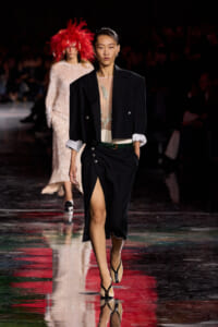 Model walks the runway in a black blazer over a sheer embroidered top and a high-slit black skirt with a green belt, wearing black strappy heels.