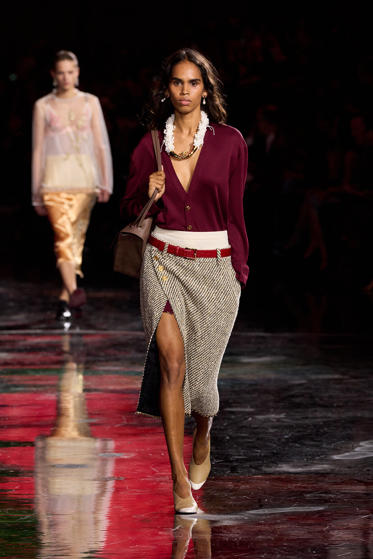 Female model walking a runway in a burgundy V-neck cardigan, white belt, and tweed wrap skirt with a front slit; carrying a brown bag and wearing a chunky necklace and beige heels.