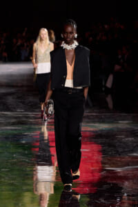 Model walks the runway in a black blazer over a peach blouse with black trousers, a white belt, a large floral necklace, and a metallic bag with chain strap.