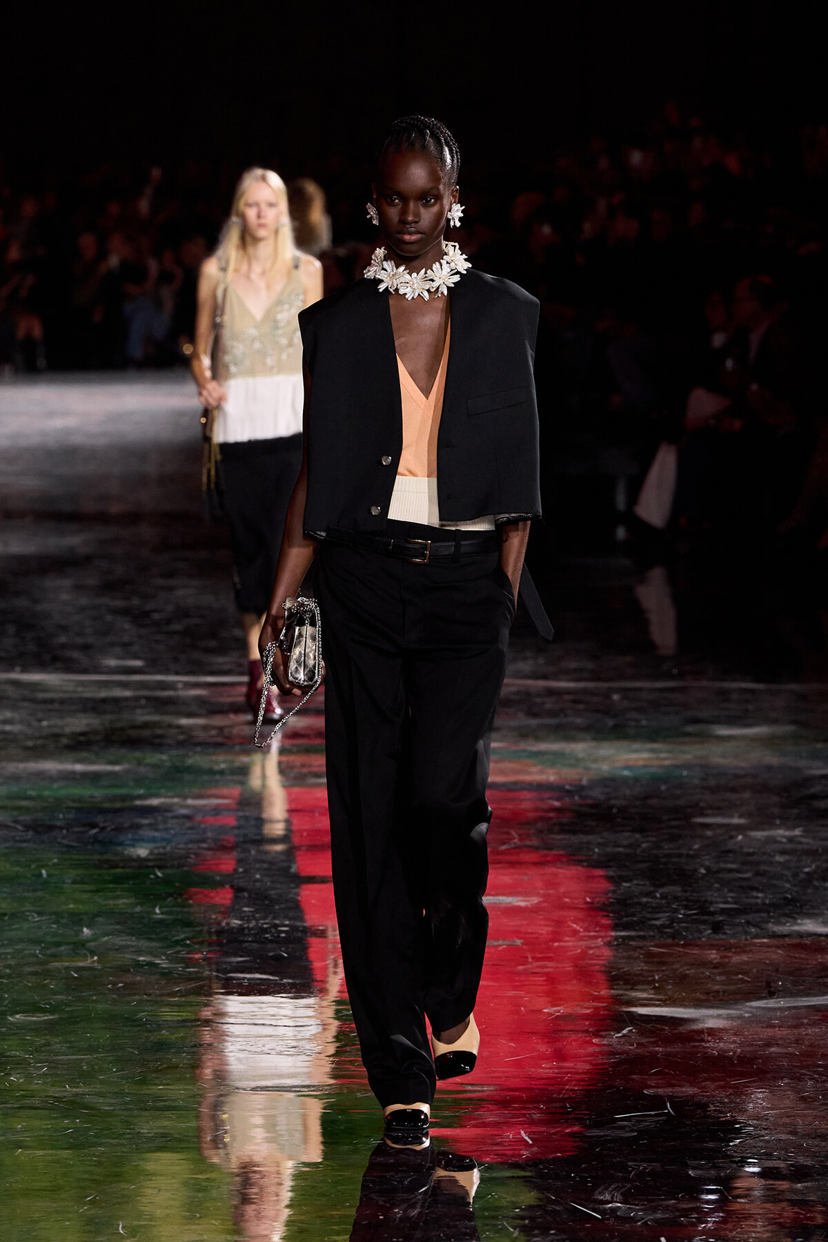 Model walks the runway in a black blazer over a peach blouse with black trousers, a white belt, a large floral necklace, and a metallic bag with chain strap.