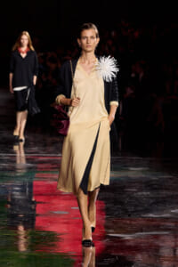Model walks the runway in a beige V-neck dress with a black overlay, navy cardigan, and a large white feather flower at the shoulder.