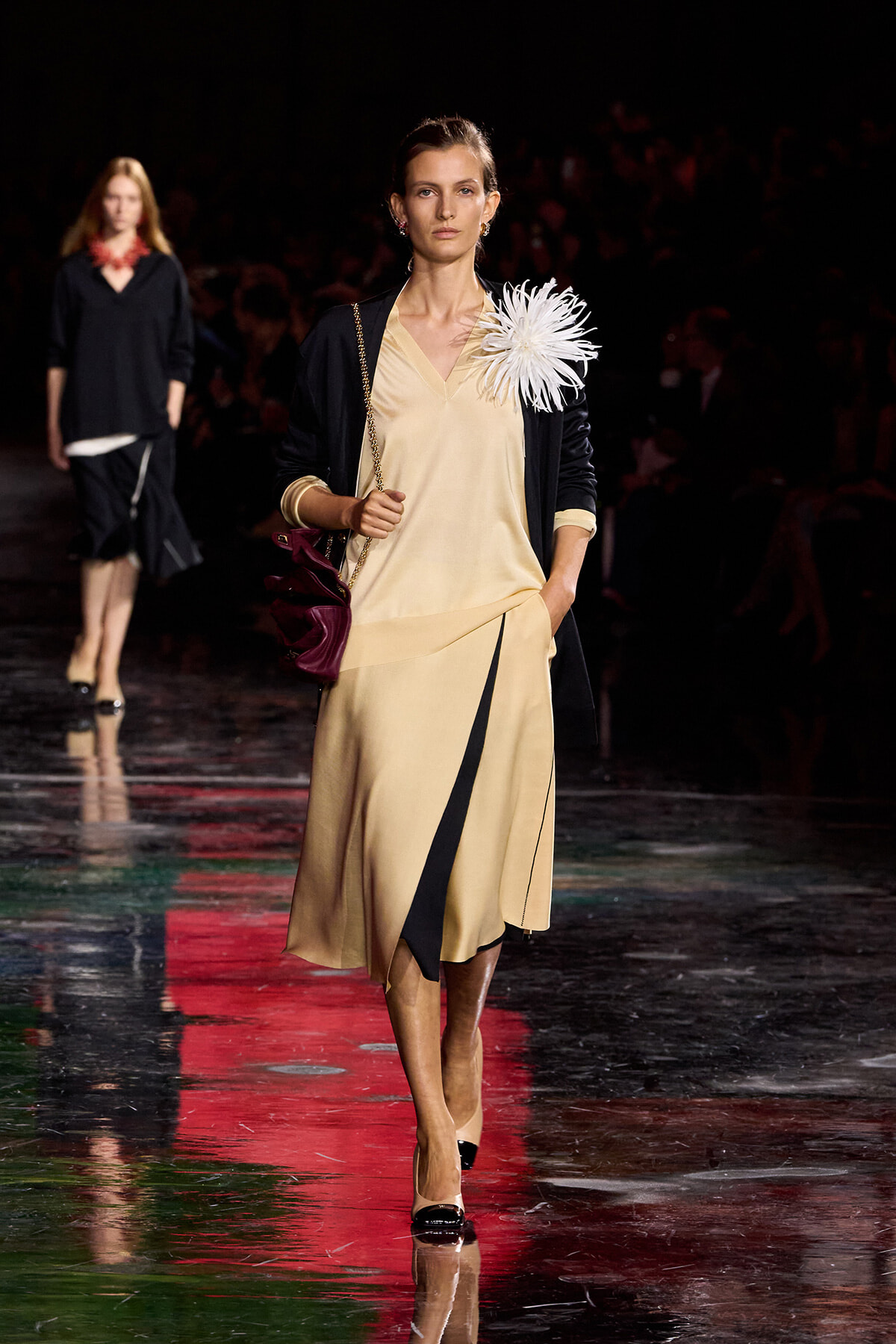 Model walks the runway in a beige V-neck dress with a black overlay, navy cardigan, and a large white feather flower at the shoulder.