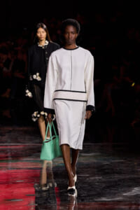 Model walks the runway in a white, long-sleeve dress with black piping and a mint-green handbag.
