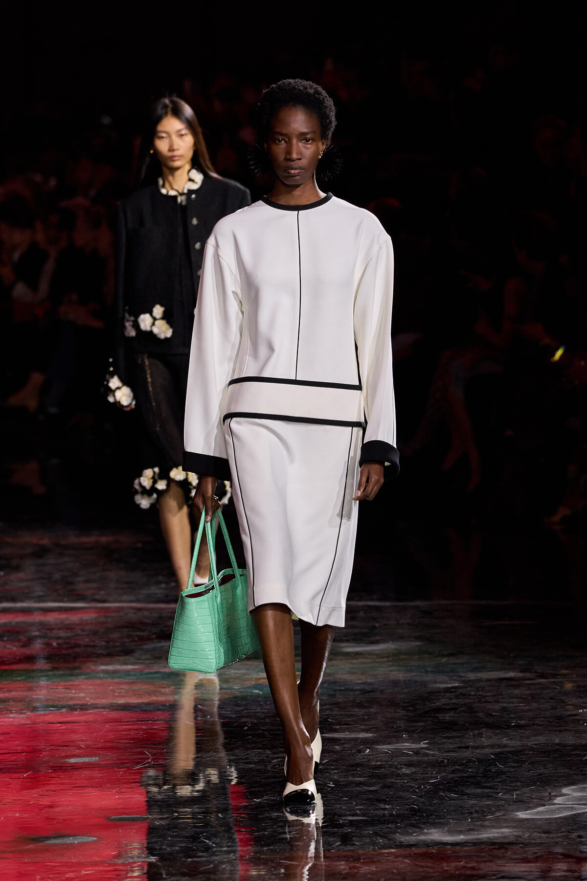 Model walks the runway in a white, long-sleeve dress with black piping and a mint-green handbag.