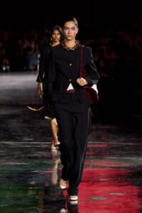 Model walks the runway in a tailored black pantsuit with gold buttons, burgundy handbag slung over her shoulder, and a chunky gold necklace under studio lighting.