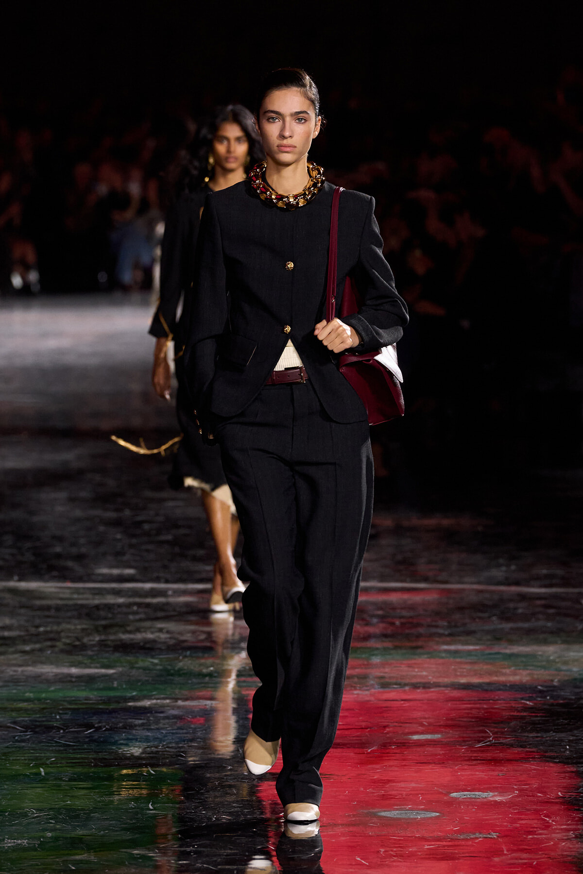 Model walks the runway in a tailored black pantsuit with gold buttons, burgundy handbag slung over her shoulder, and a chunky gold necklace under studio lighting.