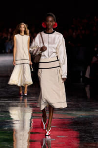 Model walks the runway in a cream satin top and beige skirt with black piping, red statement earrings, and a brown shoulder bag on a glossy reflective catwalk.