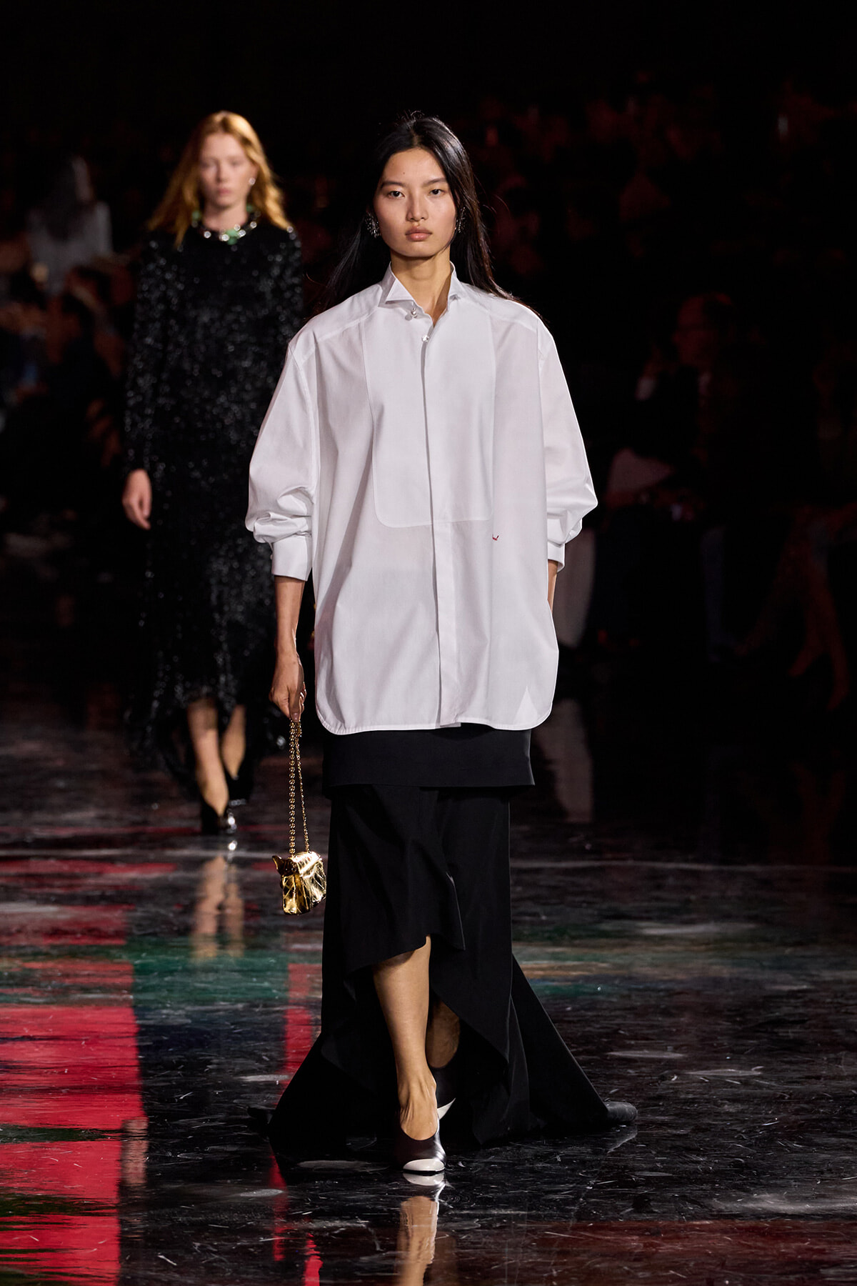 Model walks a fashion runway in an oversized white shirt and black asymmetrical skirt, carrying a small gold purse on a chain.