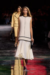 Model on a runway wearing a sleeveless ivory dress with black piping and a tiered, ruffled hem, holding a black clutch; another model in a tan coat behind.