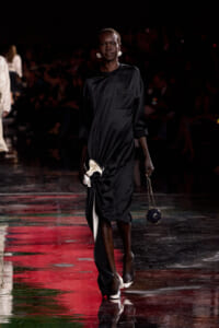 Model walks a fashion runway in a black satin midi dress, accessorized with statement earrings and a small round beaded handbag on a chain.