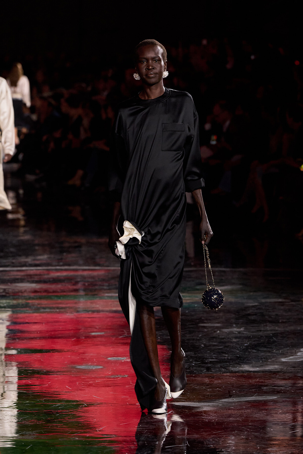 Model walks a fashion runway in a black satin midi dress, accessorized with statement earrings and a small round beaded handbag on a chain.