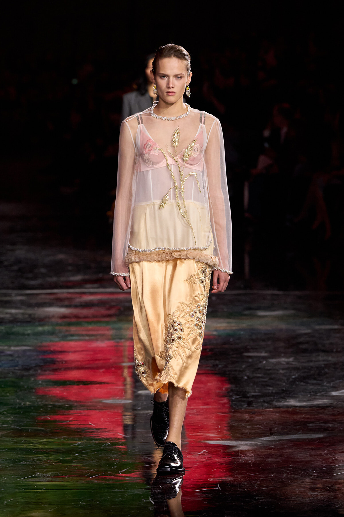 Model walking a runway in a sheer pink and cream embroidered blouse with a gold floral skirt and black loafers.