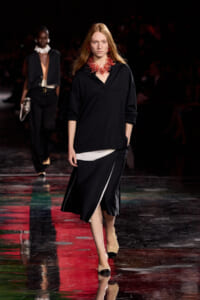 Female model walks the runway wearing a black oversized top and a black skirt with a white panel, plus a bold red coral necklace.