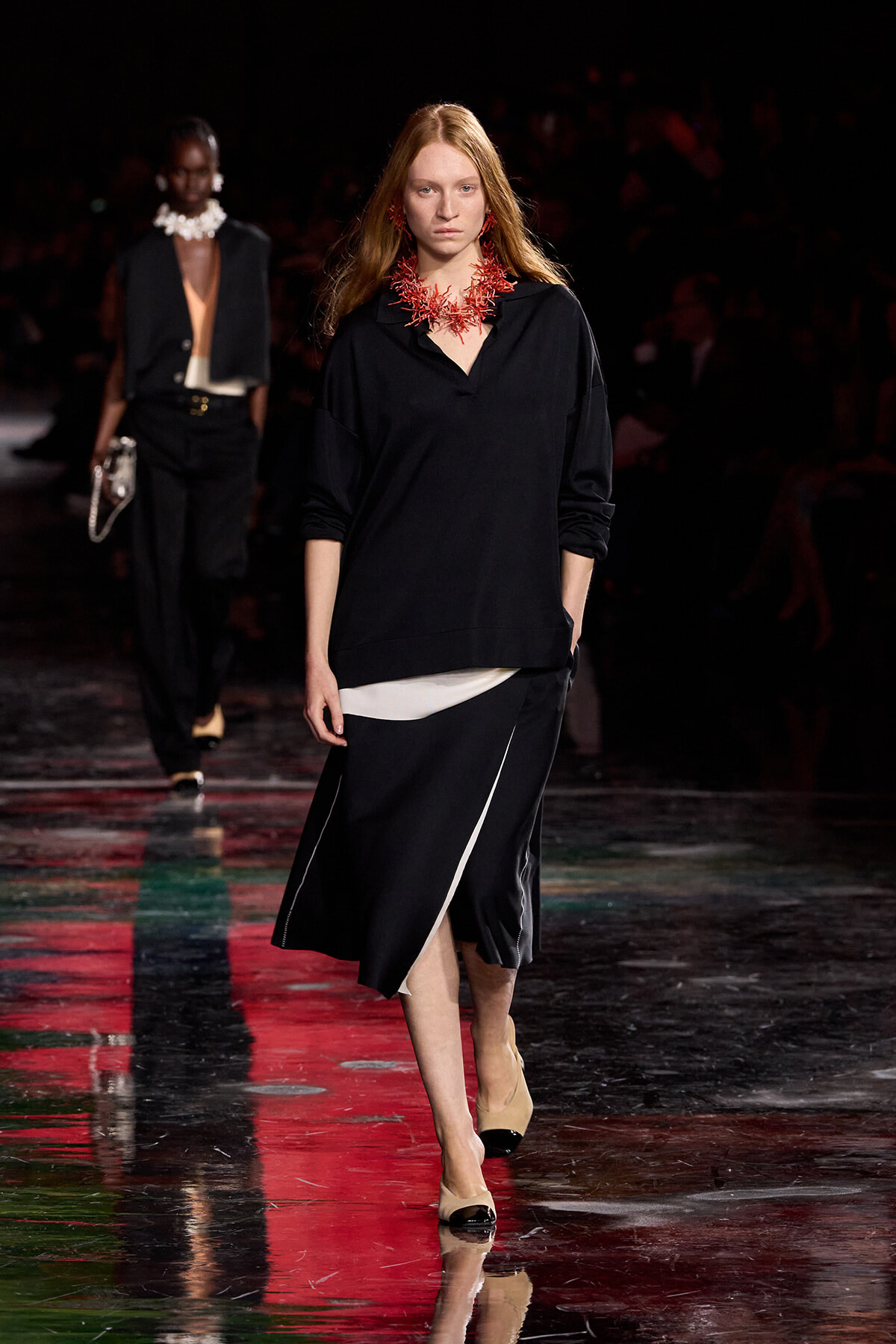 Female model walks the runway wearing a black oversized top and a black skirt with a white panel, plus a bold red coral necklace.