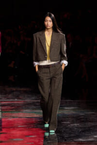 Model on a fashion runway wearing a brown tweed pantsuit with a yellow sweater, white cuffs, brown belt, and mint-green shoes.