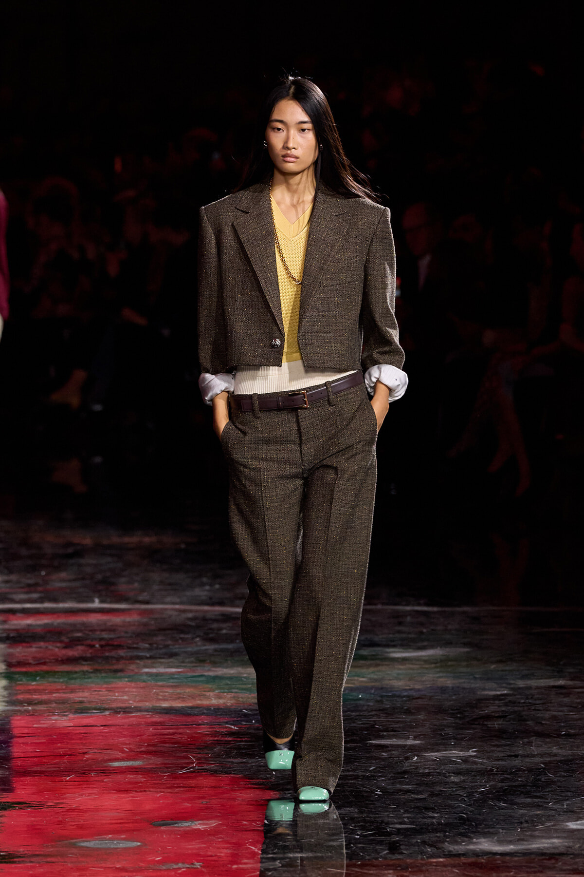 Model on a fashion runway wearing a brown tweed pantsuit with a yellow sweater, white cuffs, brown belt, and mint-green shoes.