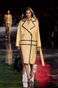 Model on a fashion runway wears a pale yellow coat with bold black piping, carrying a black handbag.