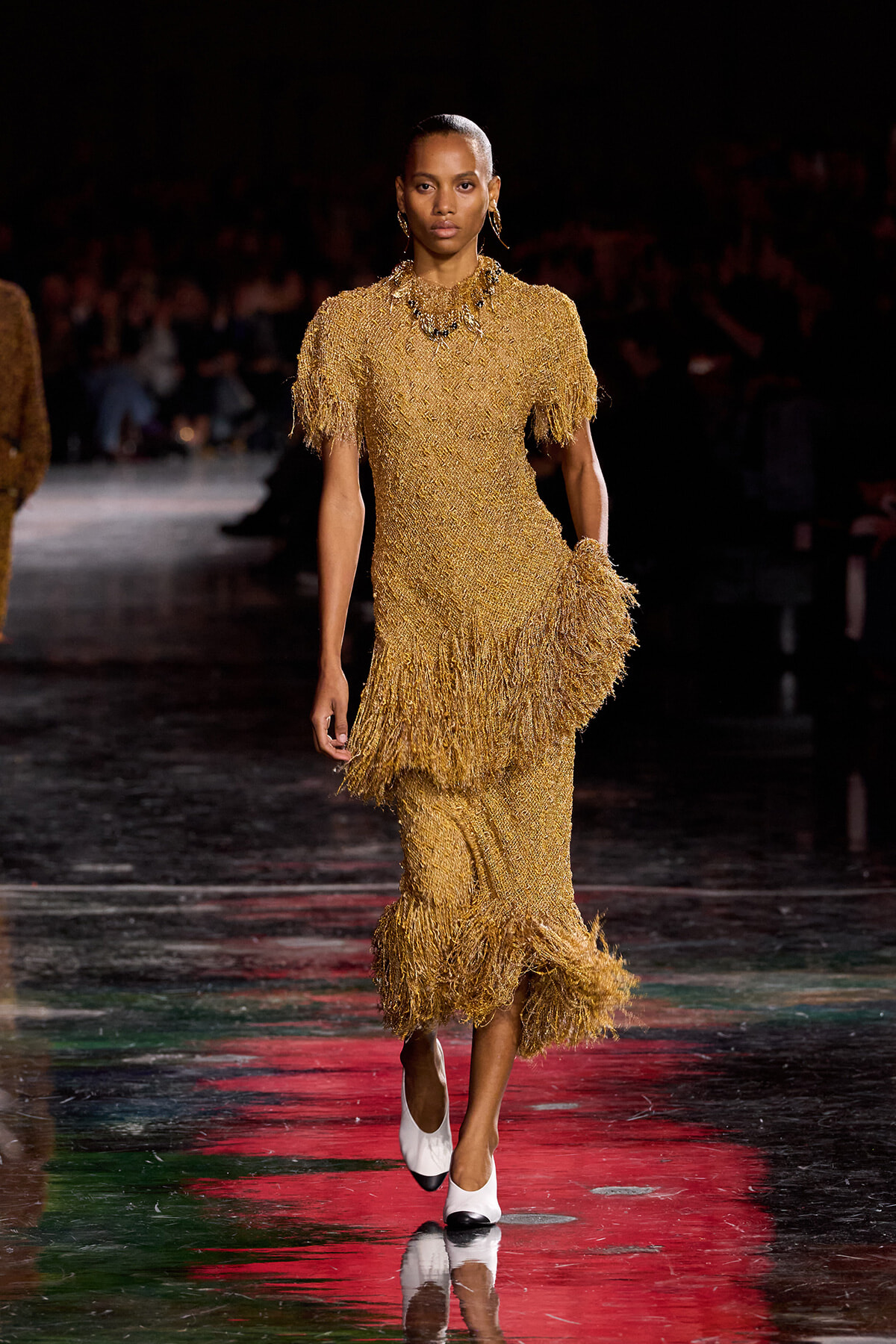 Model walks the runway in a shimmering gold fringe dress with short fringe sleeves and a hip-length fringe hem, white-and-black heels.