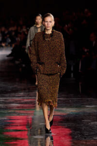 Model walking a fashion runway in a chunky brown tweed suit with fringe detailing and black heels.