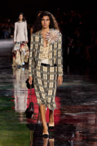 Model walks the runway in a beige plaid tweed suit with a fringed hem and a large white flower pinned at the shoulder, carrying a small burgundy handbag.