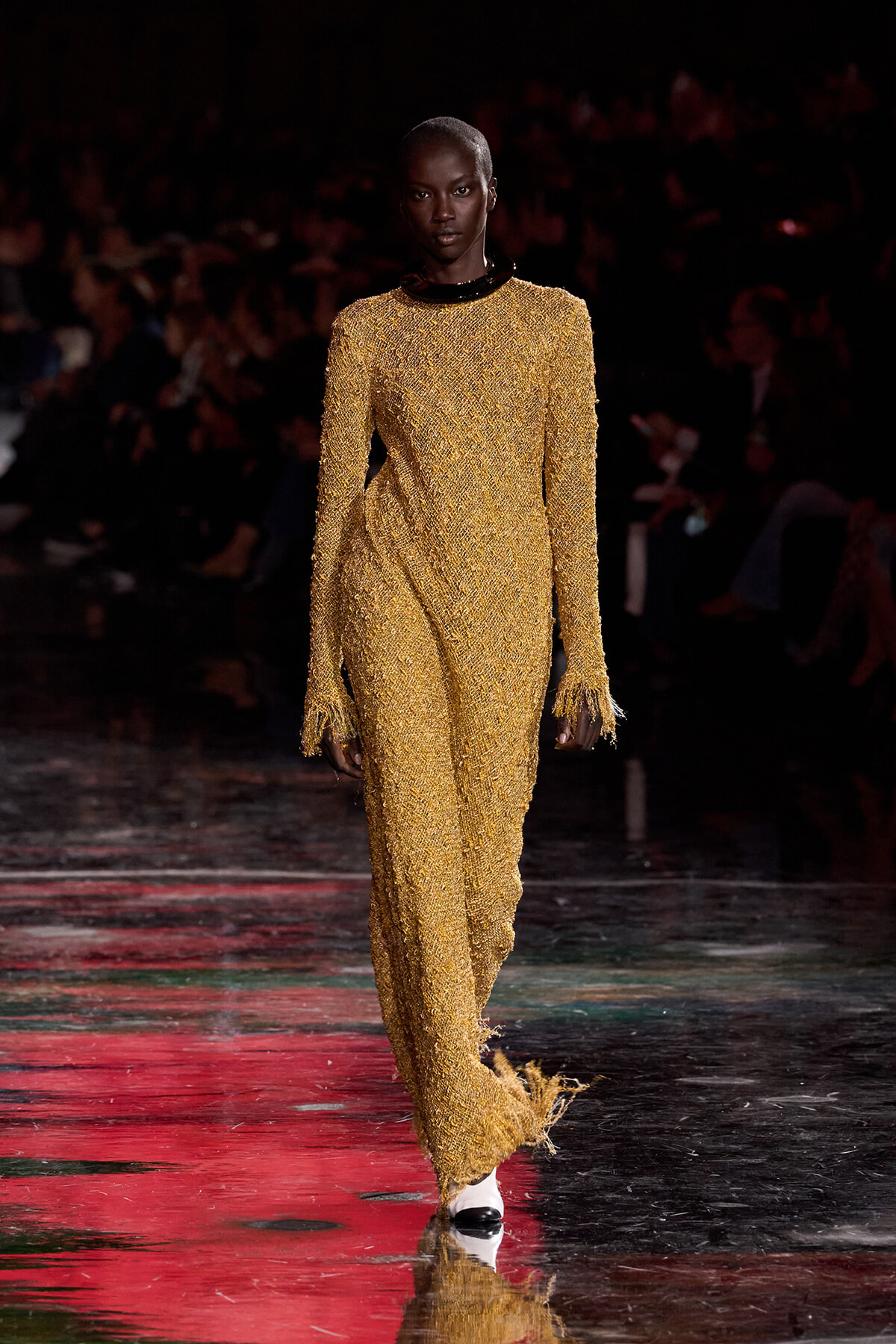 Model on a runway wearing a full-length gold beaded gown with fringed cuffs and hem, dark background audience.