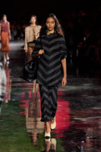 Model walking a fashion runway in a black chevron-knit tunic and matching pants, carrying a large black handbag.
