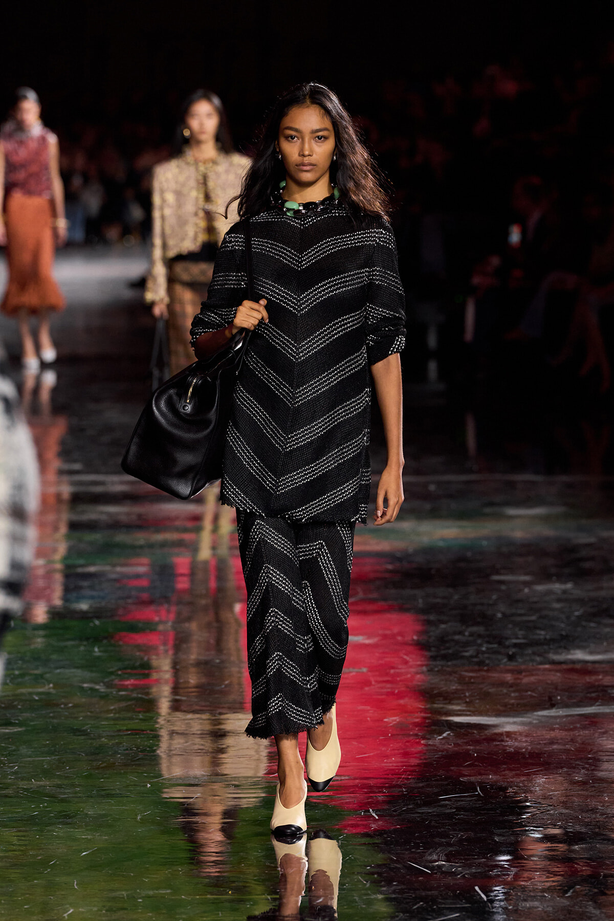 Model walking a fashion runway in a black chevron-knit tunic and matching pants, carrying a large black handbag.
