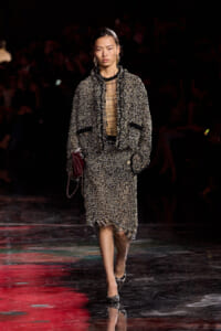 Model on a dark runway wearing a textured black-and-cream tweed suit with frayed hems, sheer blouse, gold earrings, and a burgundy clutch.