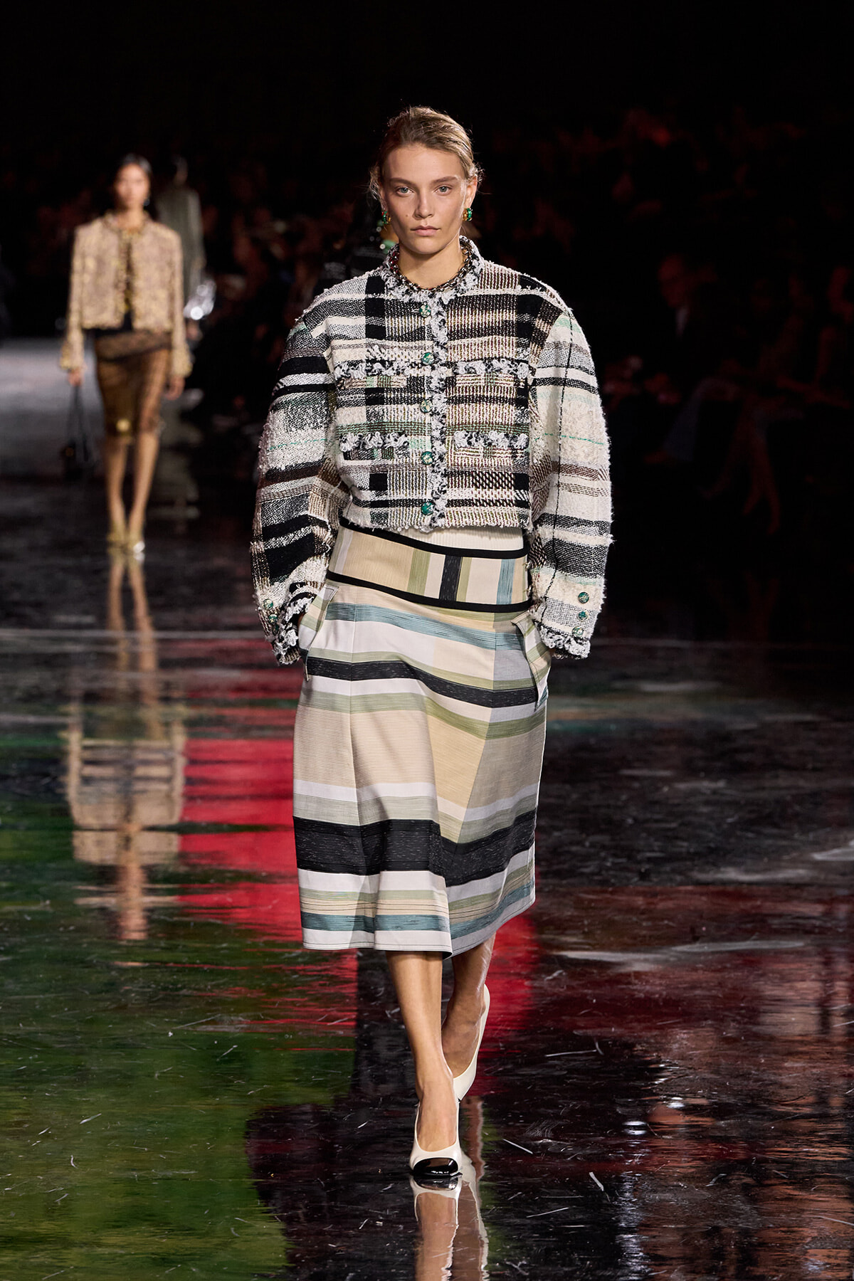 Model on the runway wearing a chunky, textured plaid jacket with green accents and a color-block striped midi skirt, white heels.